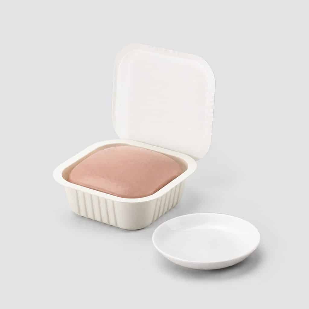 Barquette individuelle de pâtée en mousse pour chat ouverte à côté d'une assiette